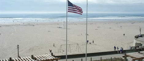 Una playa con un asta de bandera, gente disfrutando y un paseo marítimo con asientos.