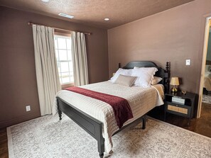 Un dormitorio con cama, mesita de noche, lámpara y ventana con cortinas.