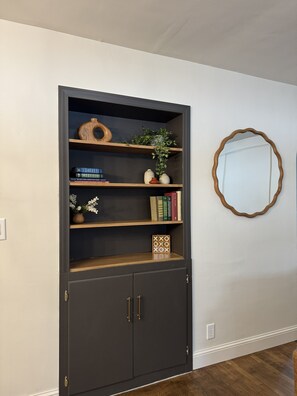 Una estantería oscura con varios estantes, algunos con objetos decorativos y libros, frente a una pared lisa con un espejo redondo.