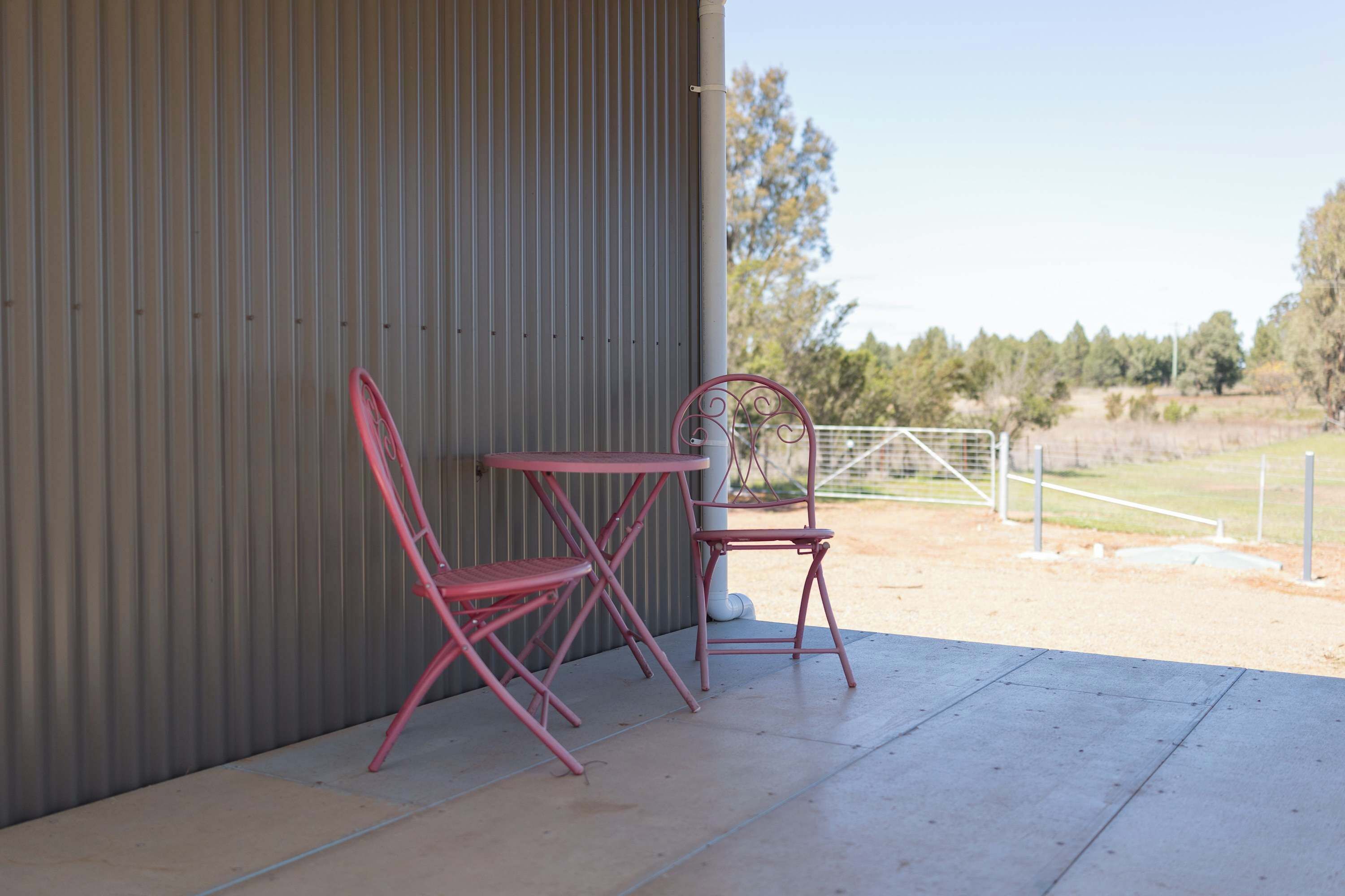 Un patio con dos sillas metálicas rojas y una mesa, un muro de lámina ondulada y vistas a un campo cercado con árboles.