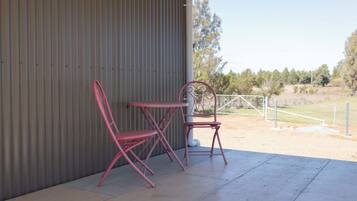 Un patio con dos sillas metálicas rojas y una mesa, un muro de lámina ondulada y vistas a un campo cercado con árboles.