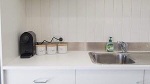 Une cuisine équipée d’une machine à café, de trois pots à épices étiquetés « sucre », « café » et « thé », d’une bouteille de liquide vaisselle Palmolive et d’un évier en acier inoxydable.