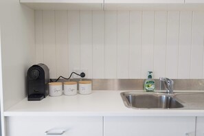 Una cocina con una cafetera, tres frascos de especias etiquetados con azúcar, café y té, una botella de detergente Palmolive y un lavamanos de acero inoxidable.