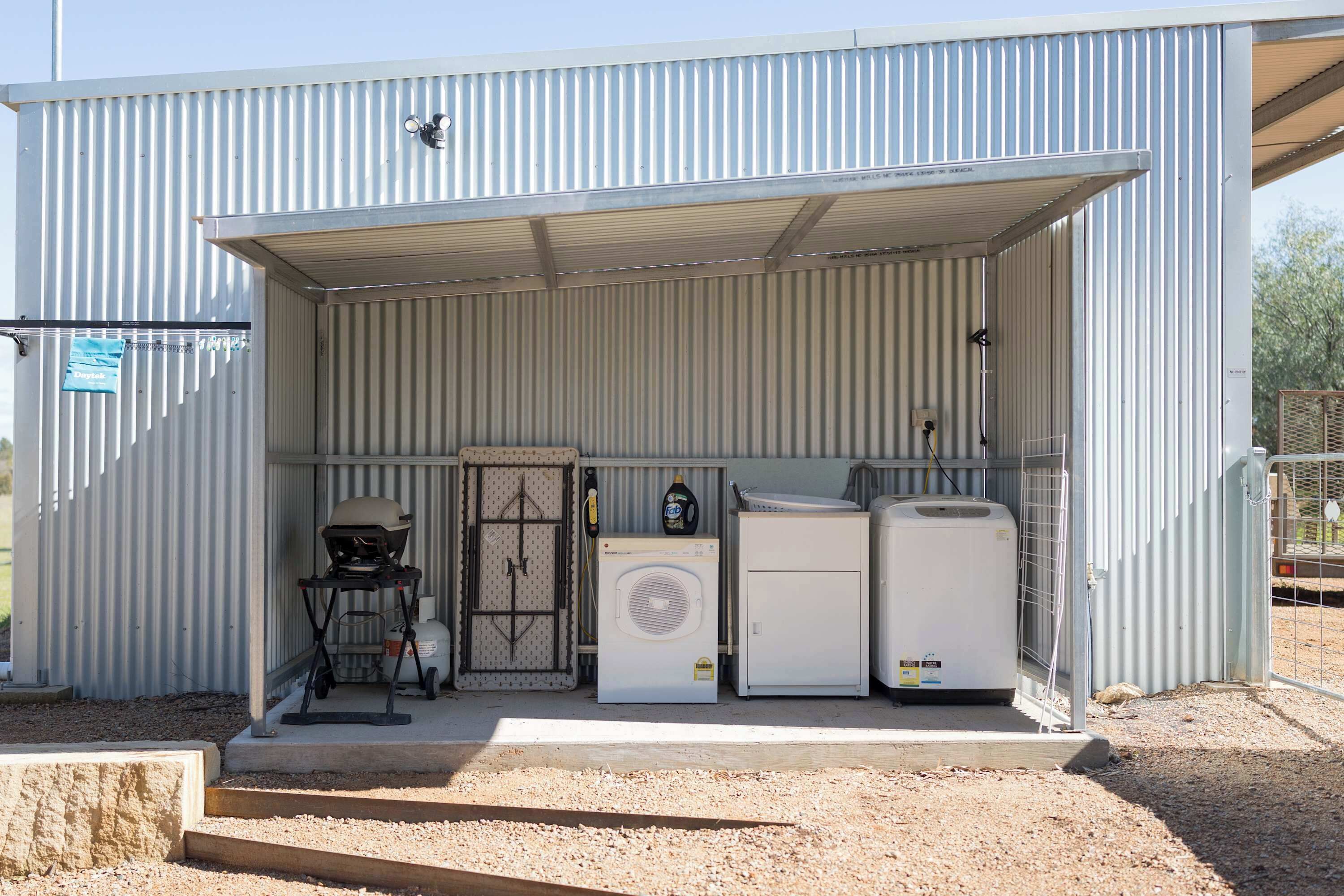 A metal building with a corrugated exterior, a covered entrance, and a gravel ground. Inside, there are two white appliances, a grill, and a water heater.