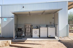 A metal building with a corrugated exterior, a covered entrance, and a gravel ground. Inside, there are two white appliances, a grill, and a water heater.