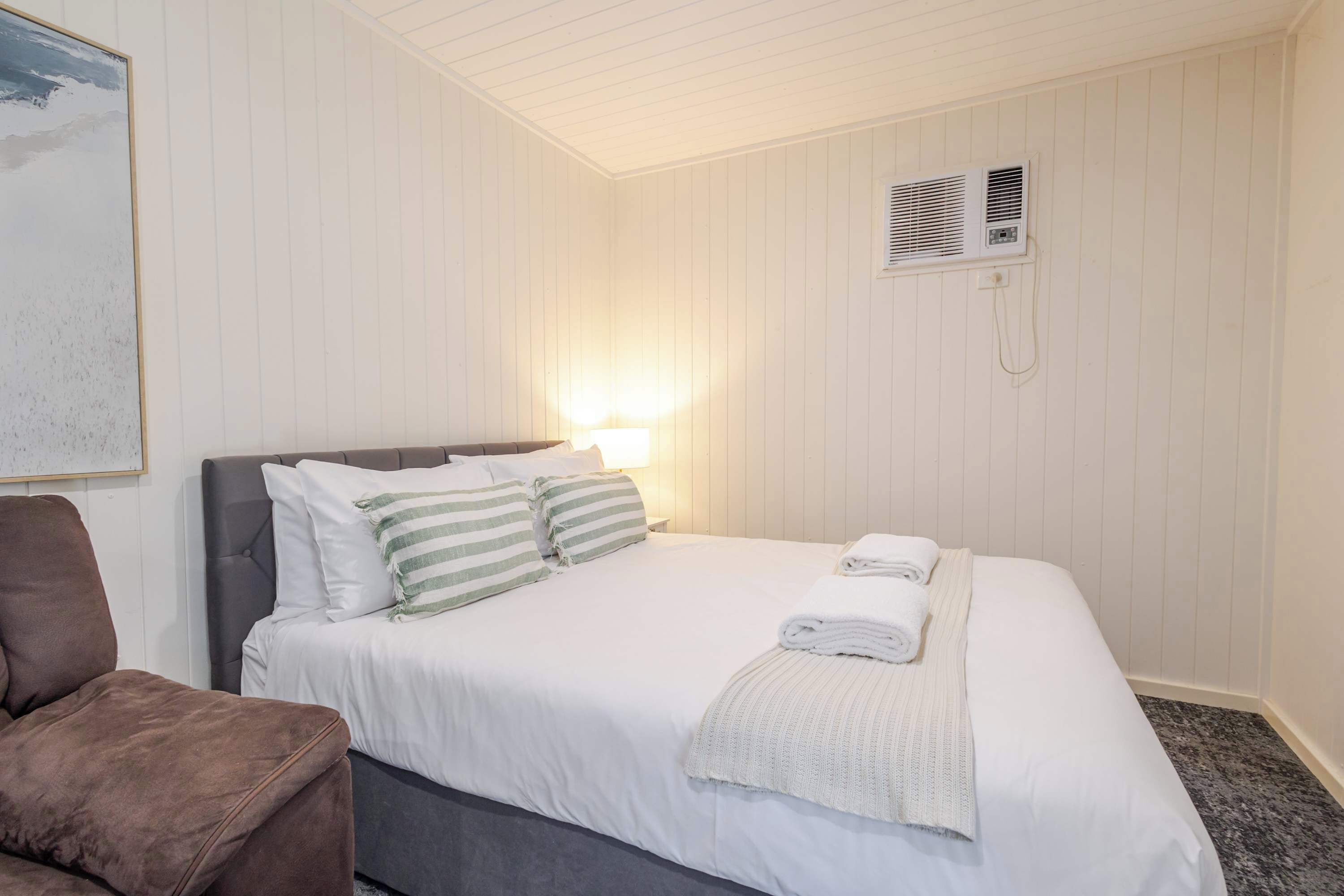 Habitación de hotel con cama, una silla, aire acondicionado de pared y un cuadro.
