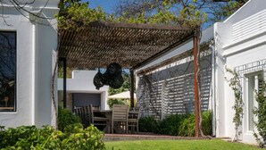 Uma varanda coberta com mesa e cadeiras, rodeada por vegetação e um edifício branco.