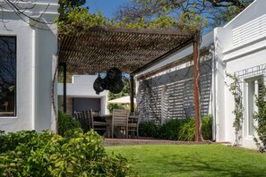 Ein überdachter Patio-Bereich mit Tisch und Stühlen, umgeben von viel Grün und einem weißen Gebäude.