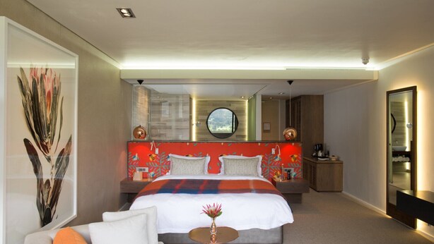 Quarto de hotel moderno com uma cama grande, um sofá, uma mesa redonda pequena e um espelho.