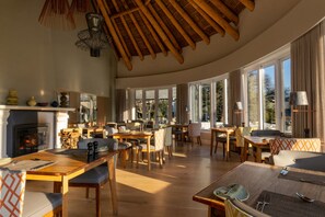 Uma sala de jantar espaçosa com mesas e cadeiras de madeira, grandes janelas com vista para o exterior e uma lareira.