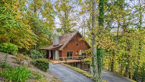 A log cabin with a porch and outdoor seating area, surrounded by trees and a driveway.
