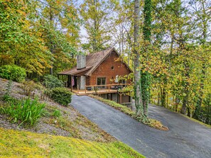 A log cabin with a porch and outdoor seating area, surrounded by trees and a driveway.