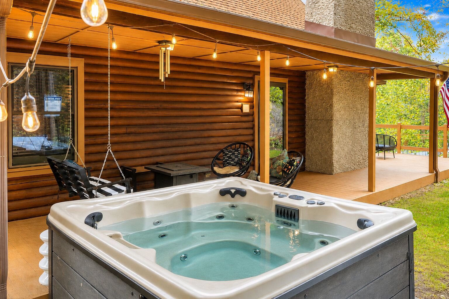 Un jacuzzi en un espacio exterior cubierto con paredes de madera, luces colgantes y mobiliario para exteriores.