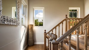 Eine Holztreppe mit Geländer, die zu einer oberen Etage führt, ein großer Spiegel mit aufwendiger Verzierung und ein Fenster mit Blick auf Grünflächen.