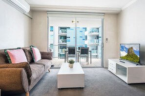 Ein modernes Wohnzimmer mit einer Couch, einem Couchtisch, einem Fernseher und einem Balkon mit Blick auf ein Gebäude.