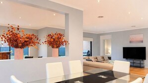 A modern living room with a glass dining table, white chairs, a sofa, and a flat-screen TV.