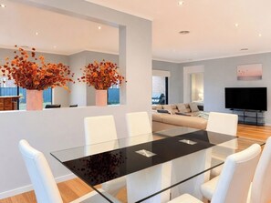 A modern living room with a glass dining table, white chairs, a sofa, and a flat-screen TV.