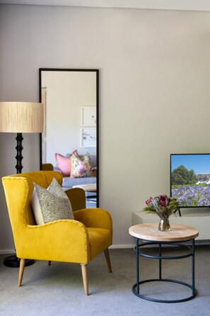 A room with a yellow armchair, a round table with a vase, a large mirror, and a television displaying a landscape. - Cap Classique Vineyard Pool Suites with Private Pool 1043 (Western Cape)