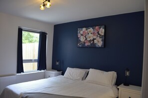 Ein Schlafzimmer mit Bett, Nachttisch, Lampe, einem Fenster mit Vorhängen und einem Gemälde an der Wand.