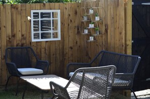 Een houten hek met een raam en een metalen plank met potplanten. Twee rieten stoelen en een klein tafeltje staan er.