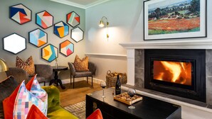 Un soggiorno con camino, un tavolino da caffè con bicchieri da vino e stuzzichini, un divano con cuscini colorati e una parete decorata con arte geometrica.