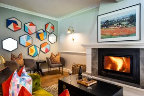Ein Wohnzimmer mit Kamin, einem Couchtisch mit Weingläsern und Snacks, einer Couch mit bunten Kissen und einer Wand, die mit geometrischer Kunst dekoriert ist.