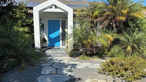 Polku johtaa talolle, jossa on sininen ovi. Taloa ympäröi vehreä kasvillisuus, ja talon edessä on kivestä tehty polku.