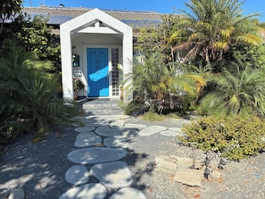 Ein Pfad führt zu einem Haus mit blauer Tür, umgeben von üppiger Bepflanzung und einem Kiesweg.
