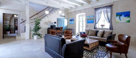 Uma sala de estar ampla com um sofá, poltronas e uma mesa de centro. Escada leva a outro nível, e uma grande janela com cortinas.
