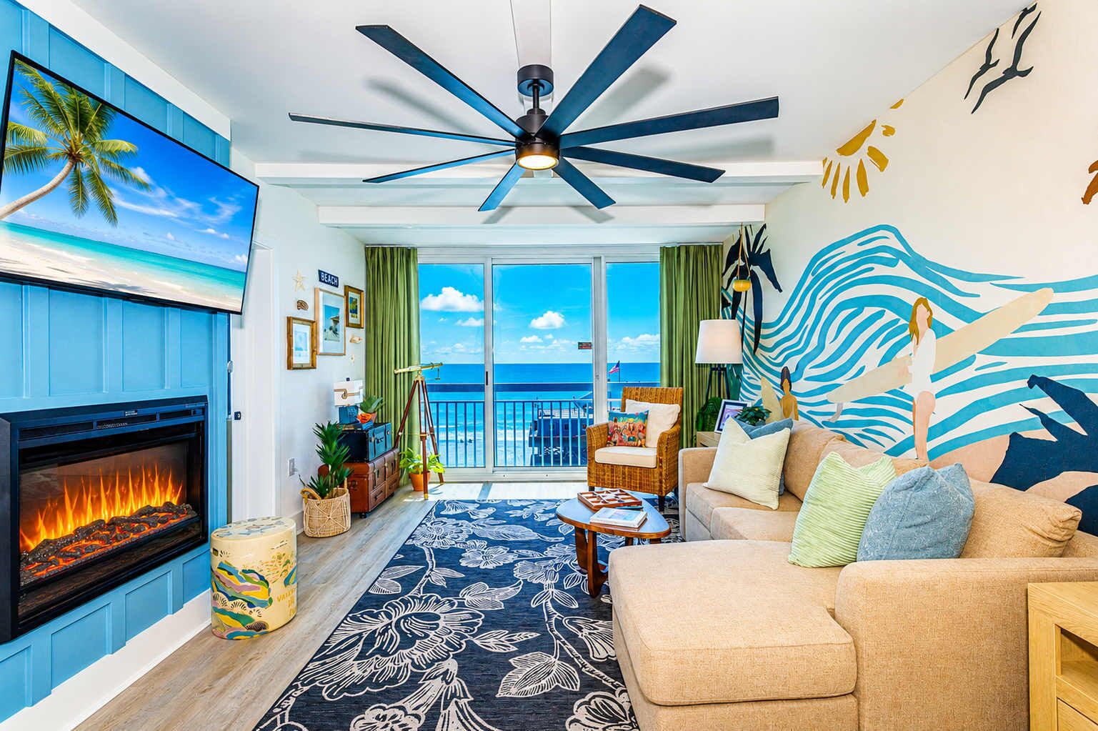 A living room with a fireplace, a large window with a view of the ocean, a ceiling fan, and a beach-themed wall mural.