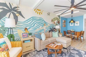A living room with a sofa, coffee table, and a wall mural. There is a ceiling fan, a lamp, and a dining area with chairs and a table. - Luxurious Coastal Oceanfront Suite! Yachtsman 601 (Myrtle Beach)