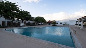 Una piscina con superficie de agua azul cristalina, rodeada por un solárium empedrado y un edificio blanco con techo de tejas rojas al fondo.
