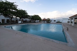 A swimming pool with a clear blue water surface, surrounded by a paved deck and a white building with a red-tiled roof in the background. - Modern 2BR 2BA Oasis in Portmore • Smart TV • AC • Free Parking (Portmore)