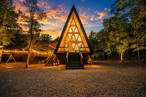 Eine dreieckige Holzhütte mit einer Treppe, die zum Eingang führt, umgeben von Bäumen und Kies.