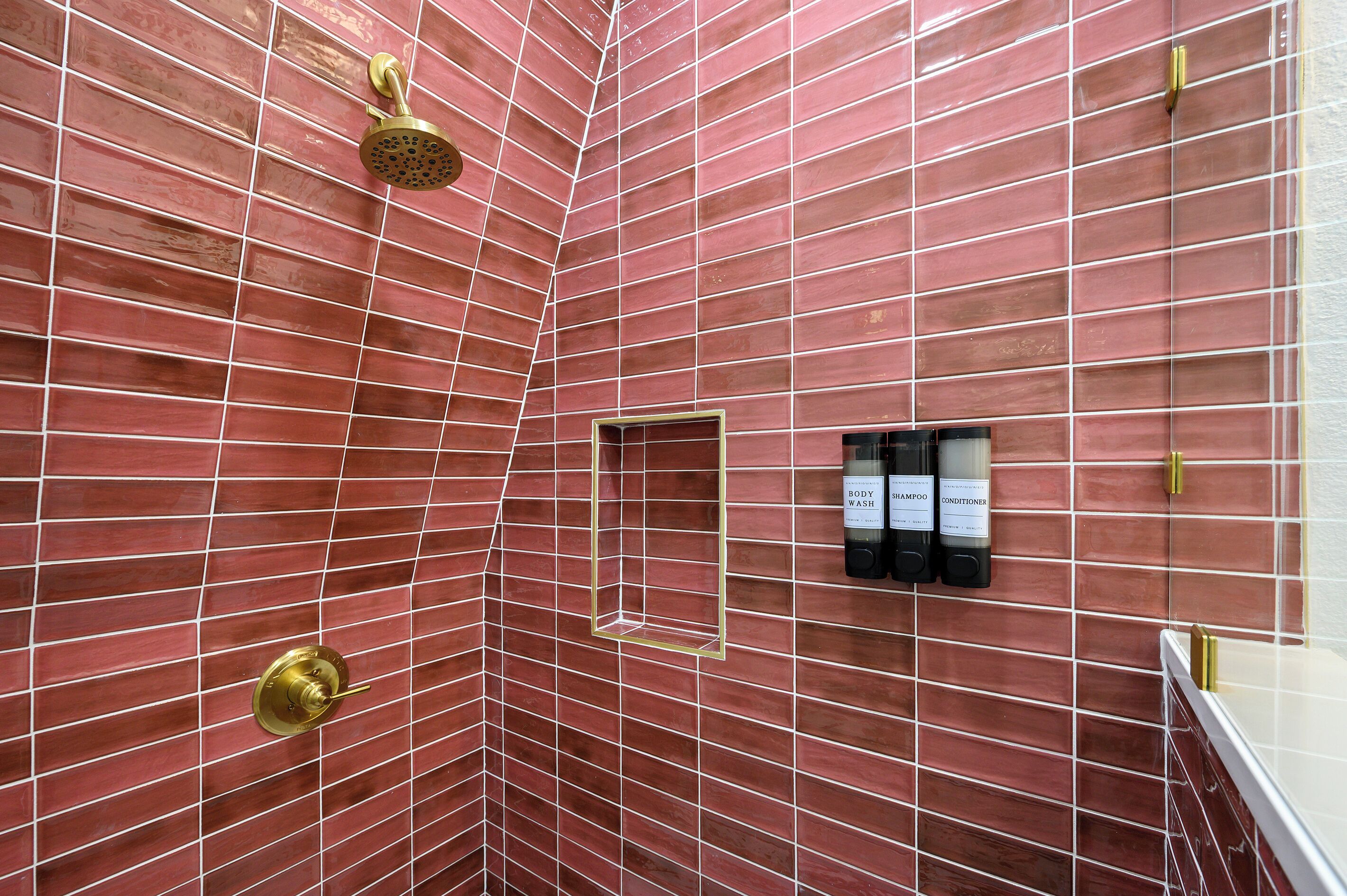 A bathroom with red-tiled walls, a showerhead, and a shelf with bottles of shampoo and conditioner.