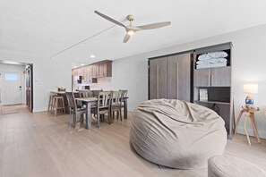 Una sala moderna con comedor, un sillón de plumas, un ventilador de techo y una cocina al fondo.