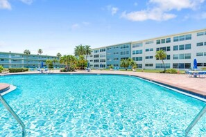 A swimming pool with clear blue water, surrounded by lounge chairs and palm trees, with a hotel building in the background. - Salty Breeze Escape|Pool|GulfView (St. Pete Beach)