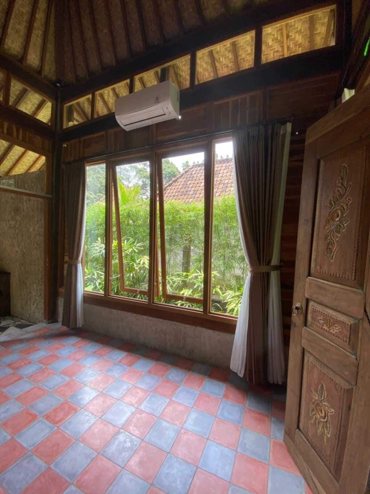 Una habitación con suelo de baldosas, ventanas amplias con cortinas, una puerta de madera con detalles decorativos y un equipo de aire acondicionado.