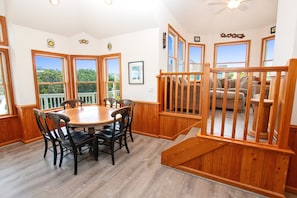 A dining area with a wooden table and chairs, a staircase with a wooden railing, and large windows with a view of greenery. - Oceanside, close to beach, Priv. Pool, Hot Tub, Dogs Welcome-  WW1033, Driftwood I (Corolla)