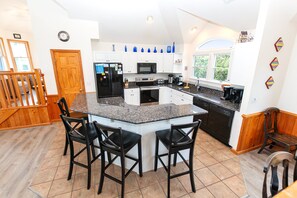 A modern kitchen with a central island, black appliances, and a dining area with chairs. - Oceanside, close to beach, Priv. Pool, Hot Tub, Dogs Welcome-  WW1033, Driftwood I (Corolla)