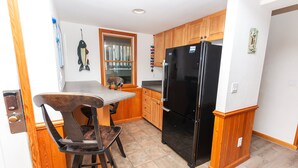 A compact kitchen with wooden cabinets, a black refrigerator, and a dining area with a table and chairs. - Oceanside, close to beach, Priv. Pool, Hot Tub, Dogs Welcome-  WW1033, Driftwood I (Corolla)