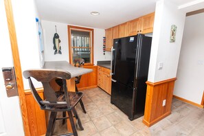 A compact kitchen with wooden cabinets, a black refrigerator, and a dining area with a table and chairs. - Oceanside, close to beach, Priv. Pool, Hot Tub, Dogs Welcome-  WW1033, Driftwood I (Corolla)