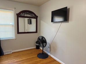 A room with a wooden floor, a standing fan, a wall-mounted TV, and a door with a mirror above it. - Hakuna Matata #6: Home for 12 People - 12 Minutes from Fort Bragg (Fayetteville)