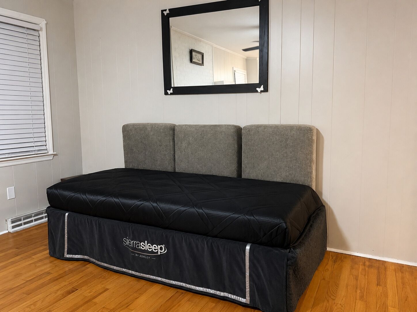 Ein Bett mit schwarzer Matratze und grauem Kopfteil in einem Zimmer mit Holzboden.