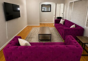 A living room with a flat-screen TV mounted on the wall, a wooden coffee table, and two purple sofas with cushions. - Hakuna Matata #6: Home for 12 People - 12 Minutes from Fort Bragg (Fayetteville)