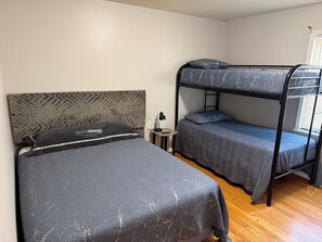 A room with two bunk beds, a small bedside table with a lamp, and a window with blinds. - Hakuna Matata #6: Home for 12 People - 12 Minutes from Fort Bragg (Fayetteville)