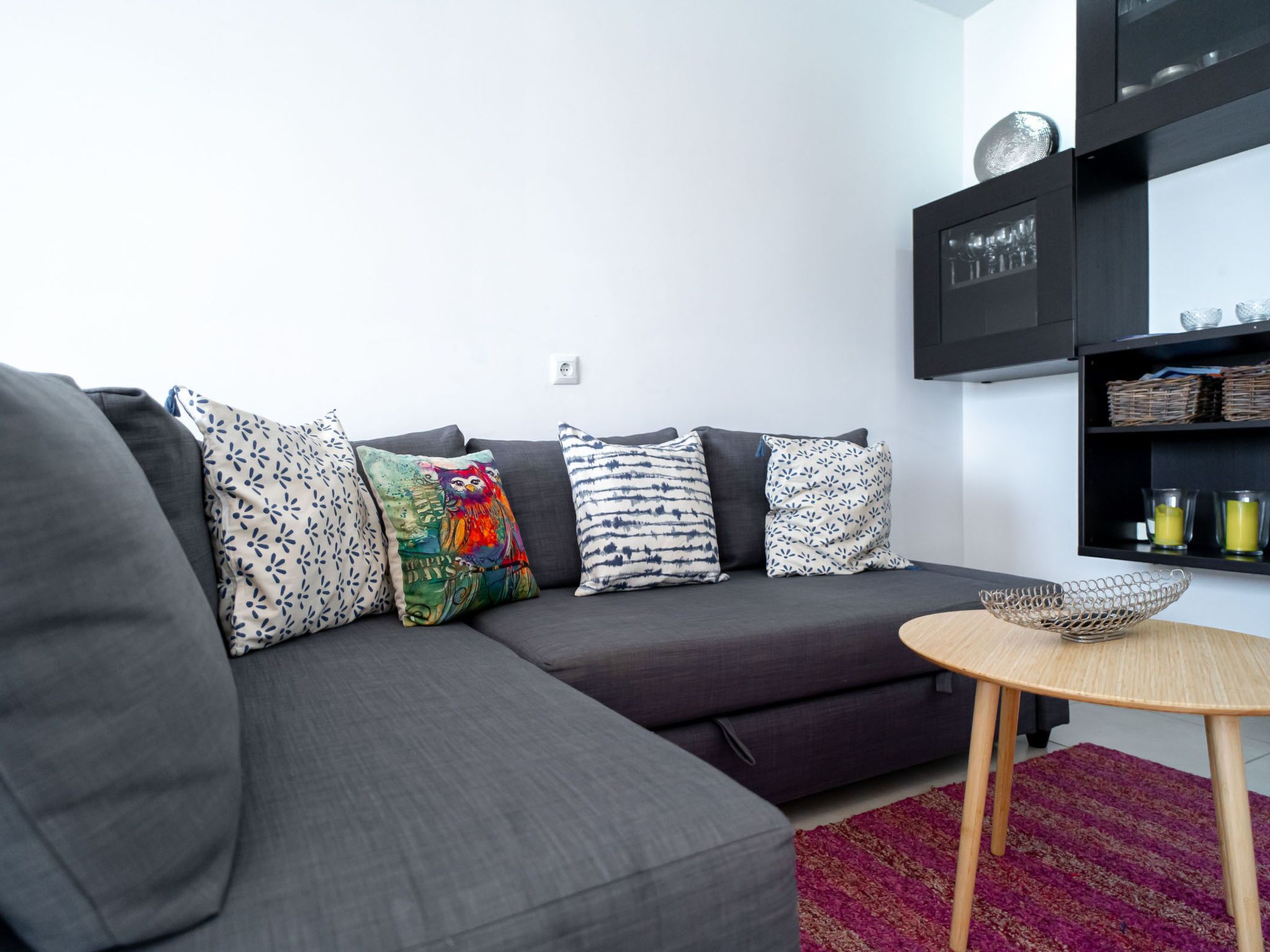 Un salon moderne avec un canapé d’angle gris, des coussins à motifs, une petite table ronde en bois et une étagère noire agrémentée d’objets décoratifs.