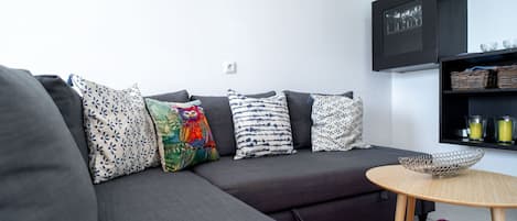 A modern living room with a grey sectional sofa, patterned cushions, a small round wooden table, and a black shelving unit with decorative items.