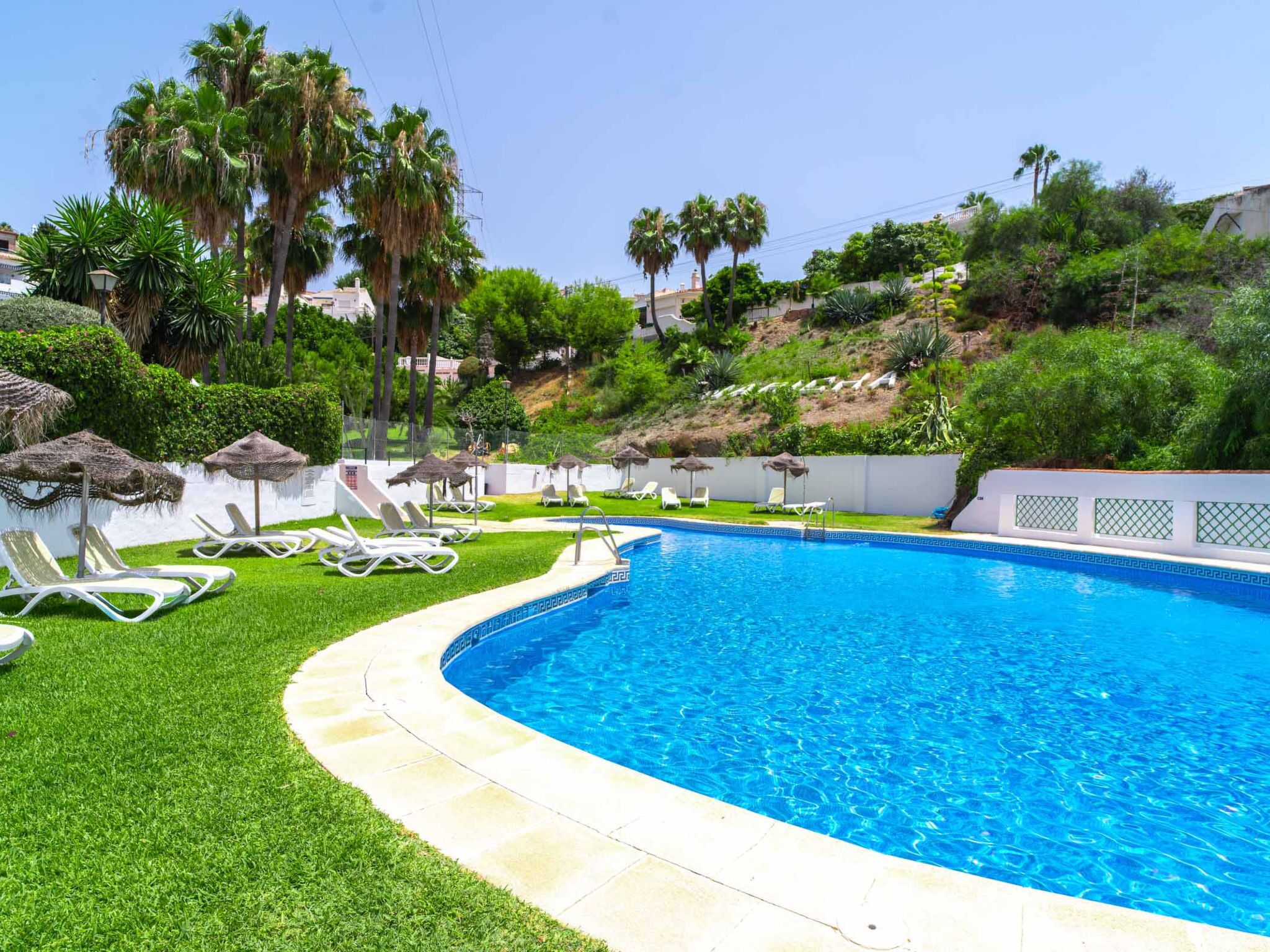 A swimming pool with a curved edge, surrounded by a white fence, and equipped with lounge chairs and umbrellas.