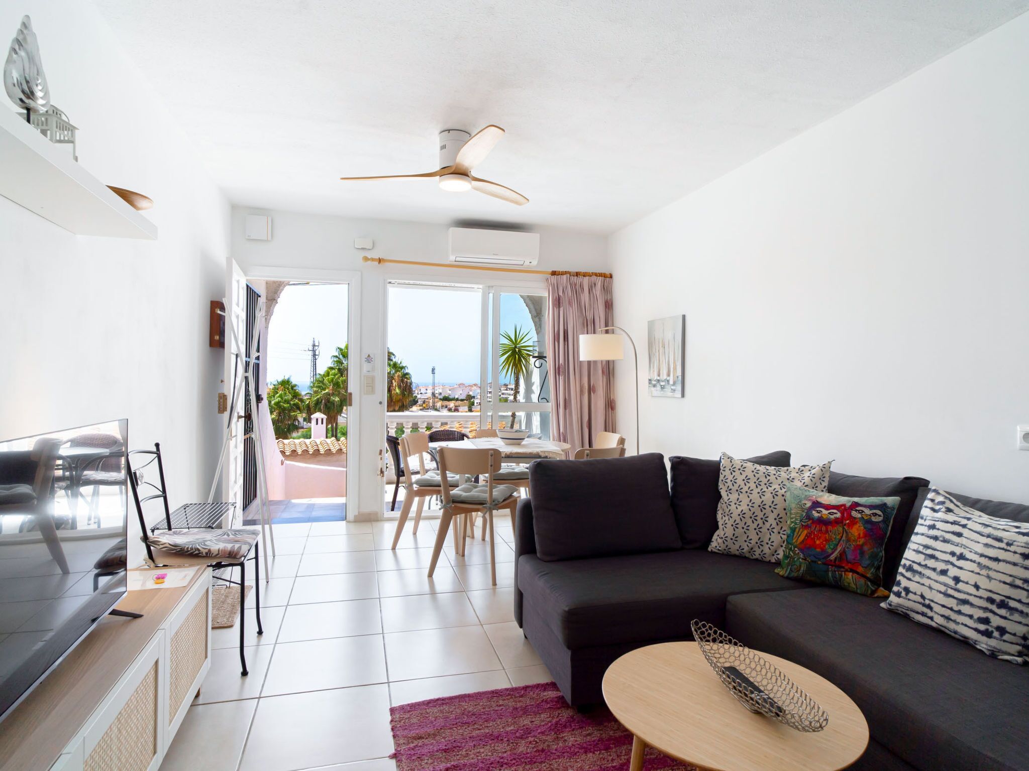 A modern living room with a sofa, a dining table, and chairs. There is a balcony with a view of the city and palm trees.